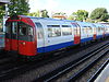 A side view of a tube train with red front. The sides of the carriages are white with a blue strip along the bottom quarter and red doors.
