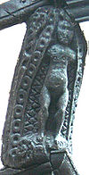 A nude female figure, lacking arms, carved in a black-painted wooden corbel. In the background are repeated decorative motifs including circles, lozenges and scrollwork.