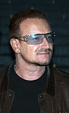 A man with facial hair wearing a leather jacket, a black shirt, an earring, and tinted glasses with a star along the frame.
