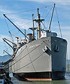 Photograph of the liberty ship Jeremiah O'Brien at dock, cranes bristling along its length. A banner on a nearby fence reads "Open", indicating its status as a museum ship.