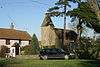 Stone church with square tower in the background partially obscured by trees. To the left is a pink painted house with red roof and in the foreground a car and grass area.