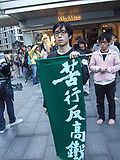 person wearing spectacles carrying a green banner with the words '苦行反高鐵' (penance against High-speed rail link) leading a group of protesters
