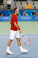 A dark-haired man is in a red shirt with white shorts and shoes and bandanna, which he is carrying his tennis racket in his right hand pointing towards the ground