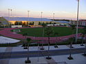 An outdoor track and field stadium with a large open grass median, and palm trees in the foreground.