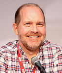 A man with closely shaven hair, and slight stubble, looking to the side slightly with his eyes, behind a microphone