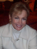 Portrait photo of a woman with blonde hair  in her late 50s smiling at the camera. She is wearing a white coat and white turtleneck underneath. (pictured in 2008)