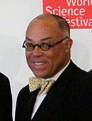 An African American male with graying hair and round eye glasses, wearing a suit with a bow tie, set against a white and red background reading World Science Festival.