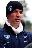 A Caucasian male wearing a blue winter jacket and a black cap both with a sports logo, a soccer ball with a wave superimposed, for the soccer team the Vancouver Whitecaps. The jacket also sports the number 27 and the subject is staring off camera.