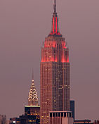 A tall, Art Deco building is shown in front of a cityscape at dusk. Lights aiming toward the top of the building glow red. The structure becomes slender as it grows taller, ending in a point at an antenna.