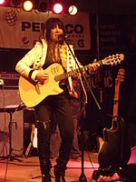 American Indian woman, wearing a white jacket and dark pants, playing a guitar and singing into a microphone.
