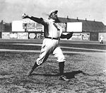 A man in a white baseball uniform and cap throws a baseball with his right hand. The shirt of his uniform has a sock-shaped icon, which reads "Boston". He is looking, and throwing, to the right of the picture.