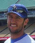 a smiling man with a LA Baseball cap and sunglasses above the caps visor smiles while wearing a stubble beard.