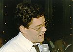 Latino man, wearing a white shirt, tie, and glasses, speaking into a microphone.