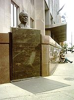 Bronze bust of a head and a plaque at a downtown street corner.