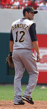 A man wearing a gray baseball uniform with the name "Sanchez" and the number "12" on the back, a black cap bearing, and a baseball glove stands on a baseball field