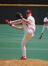 A man wearing a white baseball uniform and a red cap with a white "C" on it winds up to throw a baseball from the mound