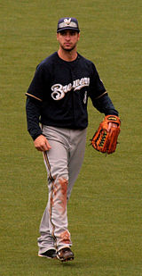 A man wearing a navy blue Brewers jersey, gray pants, navy blue cap, and outfielder's glove on his left hand walking in the outfield.