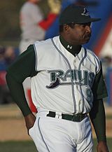 A man wearing a white baseball uniform with "Rays" written across the chest in green and a black cap stands on a baseball field