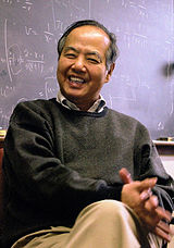 Tsung Dao Lee sitting in a chair, his legs crossed, a blackboard with mathematical equations in the background.