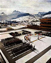 Crowds surrounding a group of flags in an open-air arena. The flags and athletes surround a rostrum. Snow-covered mountains are in the background.