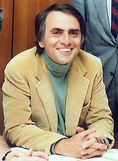 A middle-aged man sits at a desk, wearing a casual jacket and jumper. He is smiling and holding his hands together to pose for a photograph; this is physicist Carl Sagan.