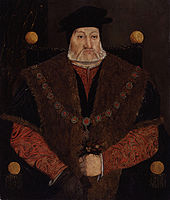 An older man with a neatly trimmed, full white beard and a composed expression wearing a red long-sleeved shirt with an intricate, wavy pattern and a black hat with short, round brim