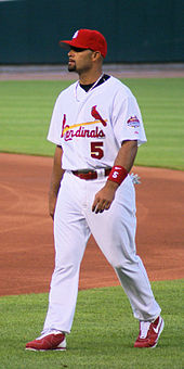 Baseball batter in a red hat, white top and white pants, standing on baseball field for the Cardinals.