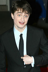 A young male with brown hair and blue eyes has his eyebrows slightly raised with his hand partly in his jacket.