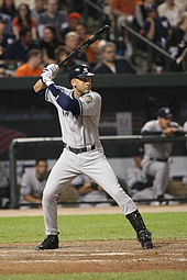 A man preparing to bat on a baseball field. He is wearing a gray jersey and pants and a dark blue batting helmet; the jersey has a blue "NEW YORK" in the front, and the helmet has a blue interlocking "N" and "Y" logo.