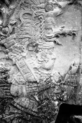 Relief sculpture of an elaborately dressed figure facing right, wearing an intricate headdress and cradling a staff in one arm.