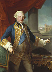 A three-quarter portrait of a young man.  His hair is light grey/blonde.  He wears pale leggings, a pale waistcoat decorated with gold lace, a large blue sash, and a blue and gold lace blazer.  His right arm rests on a chair, his left hand points to a painting behind him.