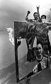 In the foreground, the canvas of a portrait of a middle-aged man and woman is ripped and burns. In the background, two men are cheering as they watch the canvas on fire.