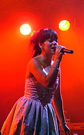  Two-thirds body shot of a young woman, with her hair pulled back and wearing a strapless dress, sings on stage into a microphone.
