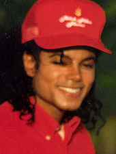 A headshot of a man wearing a red baseball cap and shirt. He has long black hair and is smiling towards the camera.