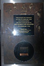 A plaque with words enclosed in a square box, underneath which is a square metal inside a circle.