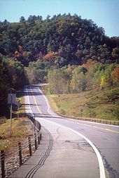 A two-lane highway in a desolate, forested area descends a hill and turns to the right and out of view ahead of a large hill in the background. The road widens to three lanes just ahead of the bend.