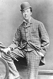 Oscar Wilde posing for a photograph, looking at the camera. He is wearing a check suit and a bowler hat. His right foot is resting on a knee high bench, and his right hand, holding gloves, is on it. The left hand is in the pocket. Oscar Wilde at Oxford.