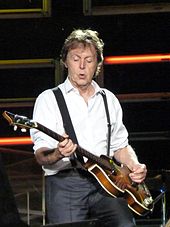A man in his sixties, wearing a white shirt and black suspenders during a concert, playing a bass guitar.