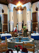 A large, open Moroccan riad that is filled with dining tables and chairs. In the center of the room is a fountain shaped like a flower with eight petals. A chandelier hangs from the ceiling, which is supported by three columns at the back. A group of people eat a meal at the table closest to the columns.