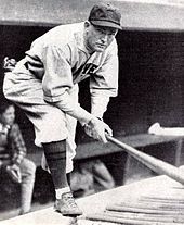 A man wearing a light-colored baseball uniform with dark socks bends over while standing in a baseball dugout.