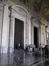 Photo shows view of vestibule with three huge doorways leading to the church's interior. The doors are framed by columns and have pediments. The floor is of inlaid marble. The nearest doorway is closed by two huge ancient bronze doors. A group listens to a tour guide while one woman examines the doors.