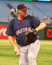 Boston pitcher, in black hat, navy blue top and grey pants, delivering a practice pitch for the Red Sox the day before a game. Nearly empty bleachers are visible behind him.