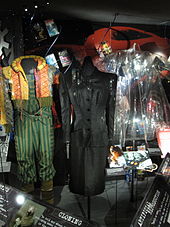 A photograph showing three costumes and some smaller items from the film. The left-hand costume is brightly coloured with striped trousers and an orange and yellow coat with a diamond pattern. The middle costume is a long black dress and the right-hand is a plastic see-through short coat.
