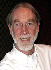 A man grins widely. He wears a white dress shirt and has a full grey beard.