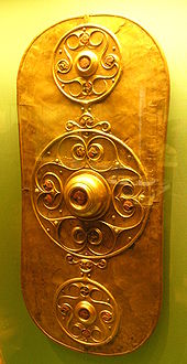 Golden shield dominated by three circular decorations. The larger central decoration has a large boss surrounded by a curlicue pattern. The smaller decorations above and below it are similar.