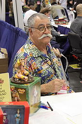 A man with thinning, greying hair and a thick, white mustache sits behind a table. He wears a colourful shirt.  Pens and action figures of his works are on the table in front of him.