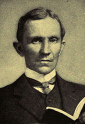  Man, probably mid-forties, dark hair, clean shaven, wearing a high collar with tie, looking staright ahead. He is holding an open book