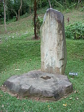 A tall plain standing stone behind a cog-shaped flat stone, in a grassy area with a jungle-covered mound rising behind.