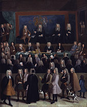 A rectangle picture of a courtroom.  Dozens of men in a courtroom in 1750s era court suits and wigs.  A blue wall at the back contains of coat of arms.  On a raised stage at the back are four men.  Several onlookers, some with dogs and children, pass by on a sidewalk looking in on the court proceedings.