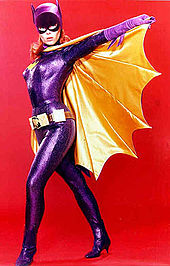 Yvonne Craig poses in the Batgirl costume from the television show, holding the cape open. The costume consists of a purple bodysuit, cowl, gloves, boots, a yellow cape and belt.